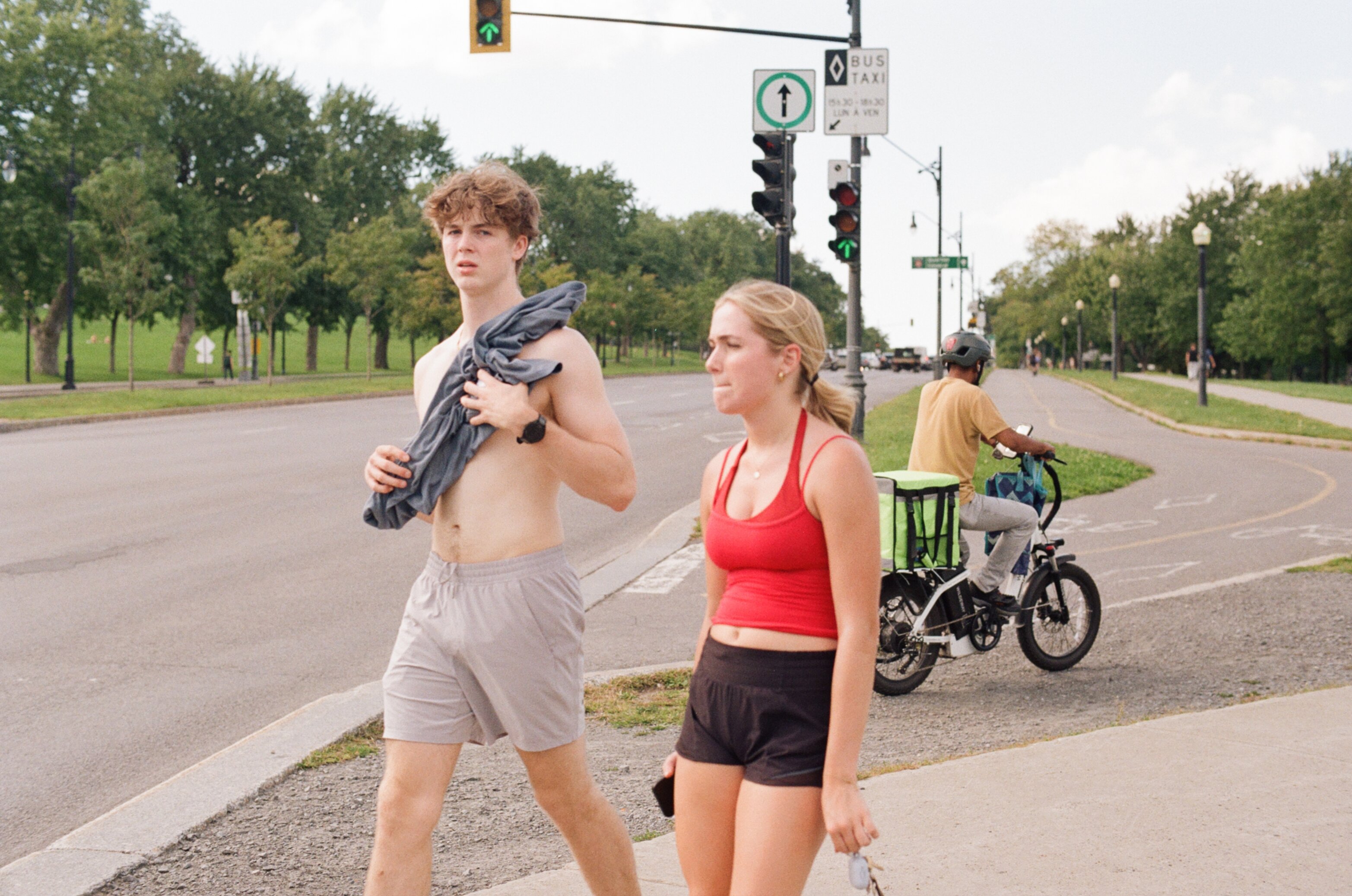 A woman and a shirtless man holding his shirt wait together to cross the street