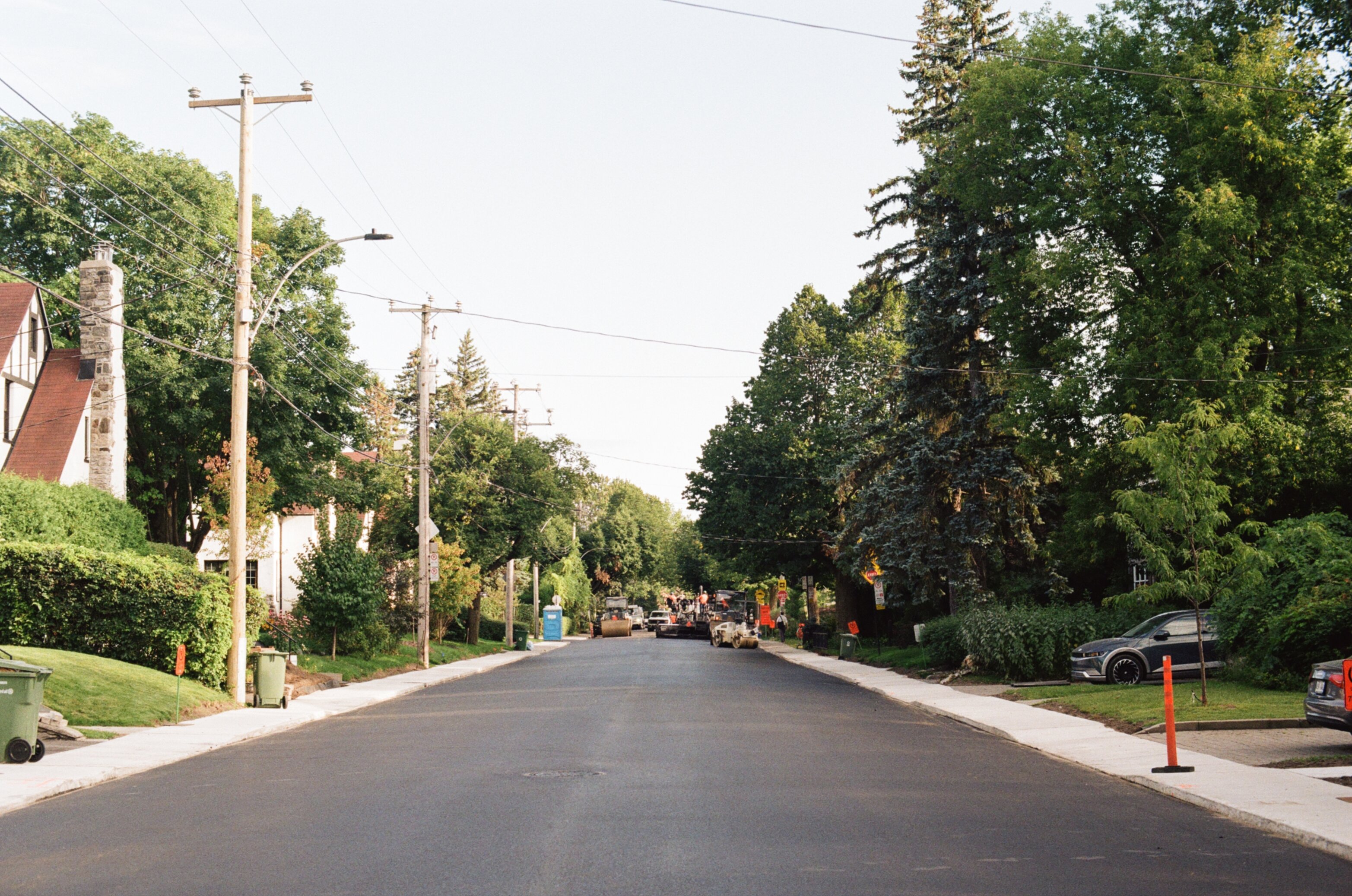 Smooth, dark freshly paved street lined with trees with steamrollers and other construction vehicles in the distance