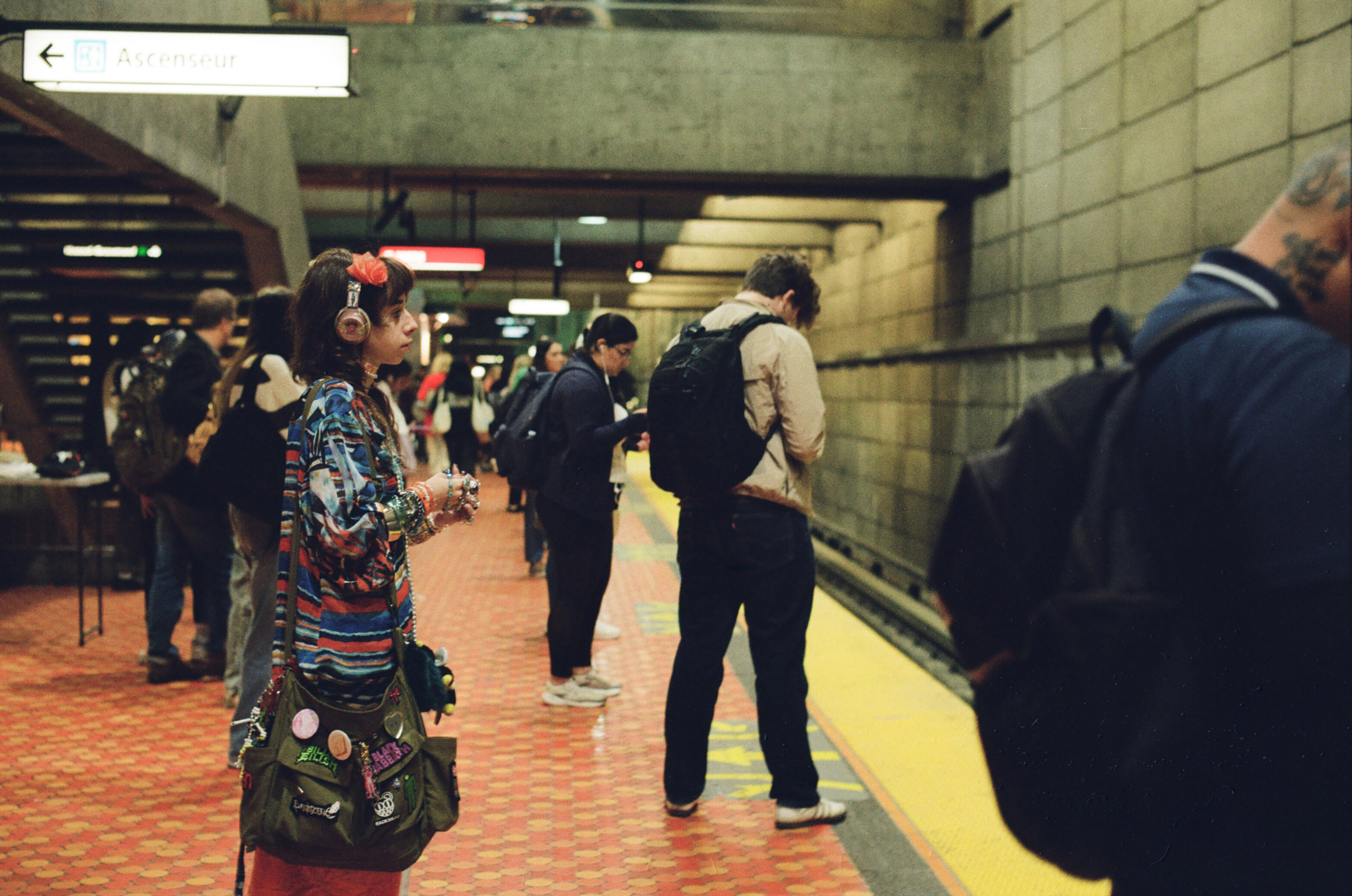 Woman with colourful clothes, headphones, headband, bracelets, and bag waiting with other passengers on the subway platform