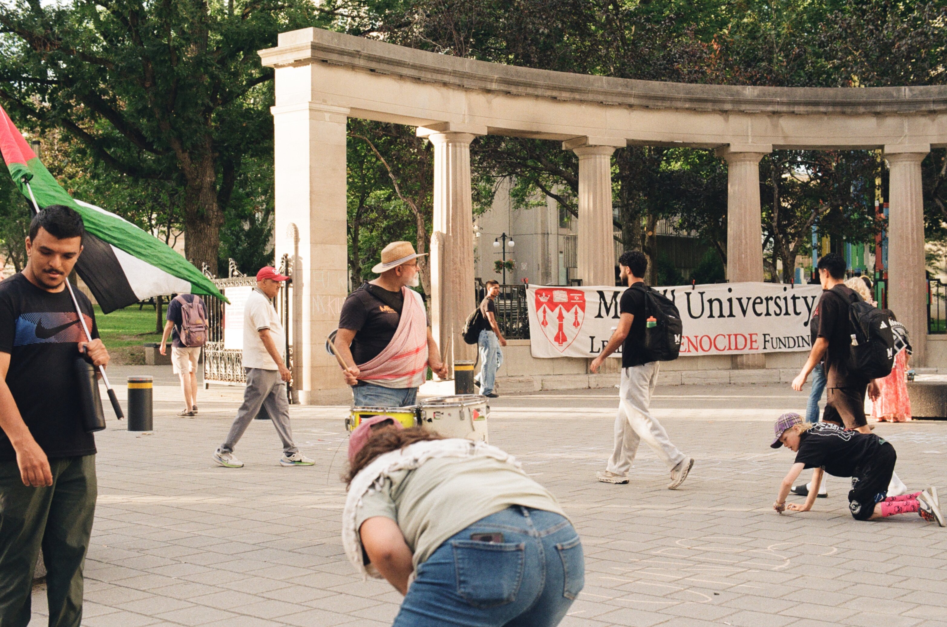A man on the left holding a Palestinian flag, a man in the middle drumming, and a woman and girl writing in chalk on the pavement with a sign in the background that says 'McGill University Leads in GENOCIDE Funding'