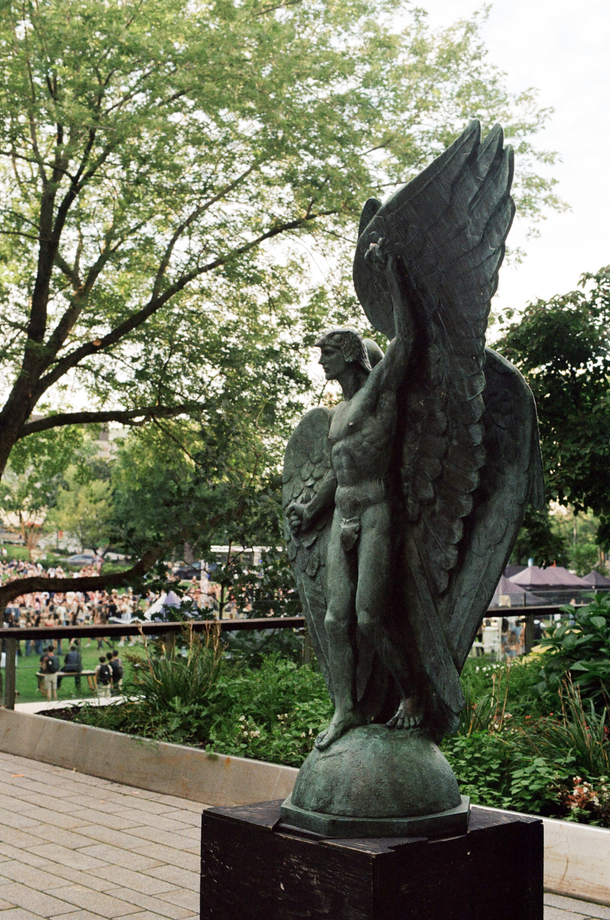 Weathered green bronze statue of a man with wings with a crowd of students visible through the trees in the background