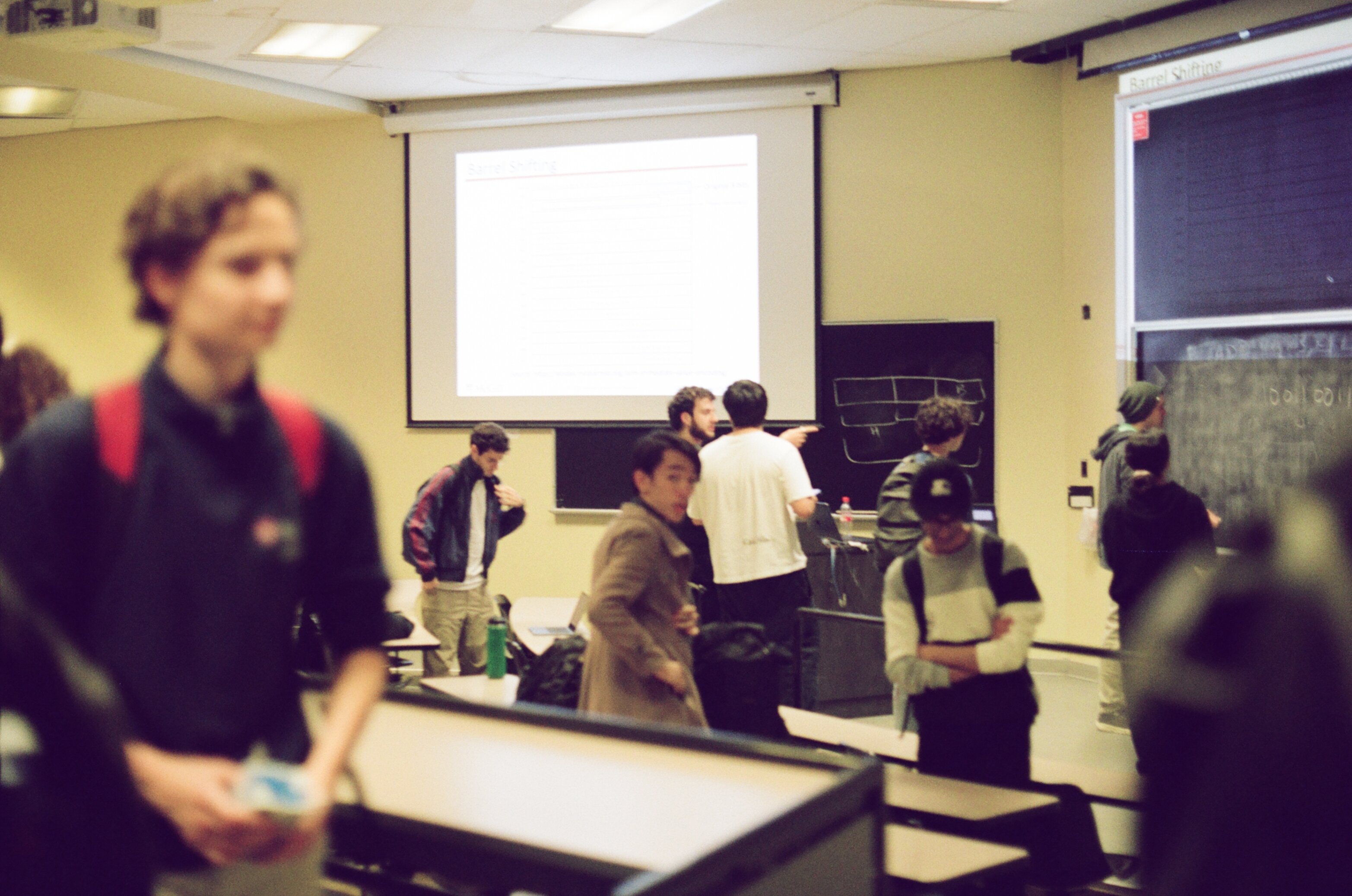 Students getting up to leave a classroom, one male student writing on a blackboard in the background