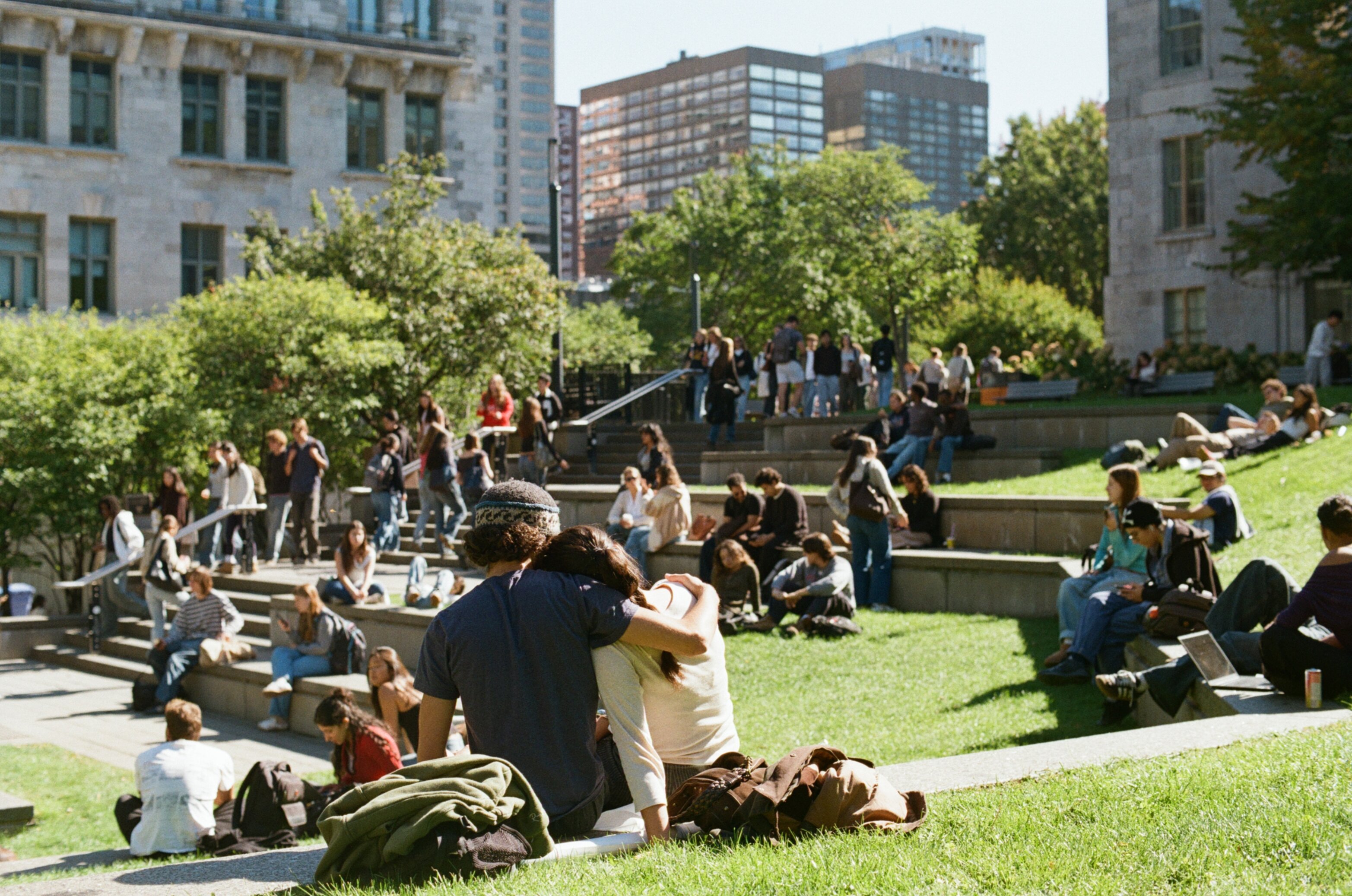 A female student rests her head on a male student's shoulder while they sit on a step in a grass field with other students sitting in the background