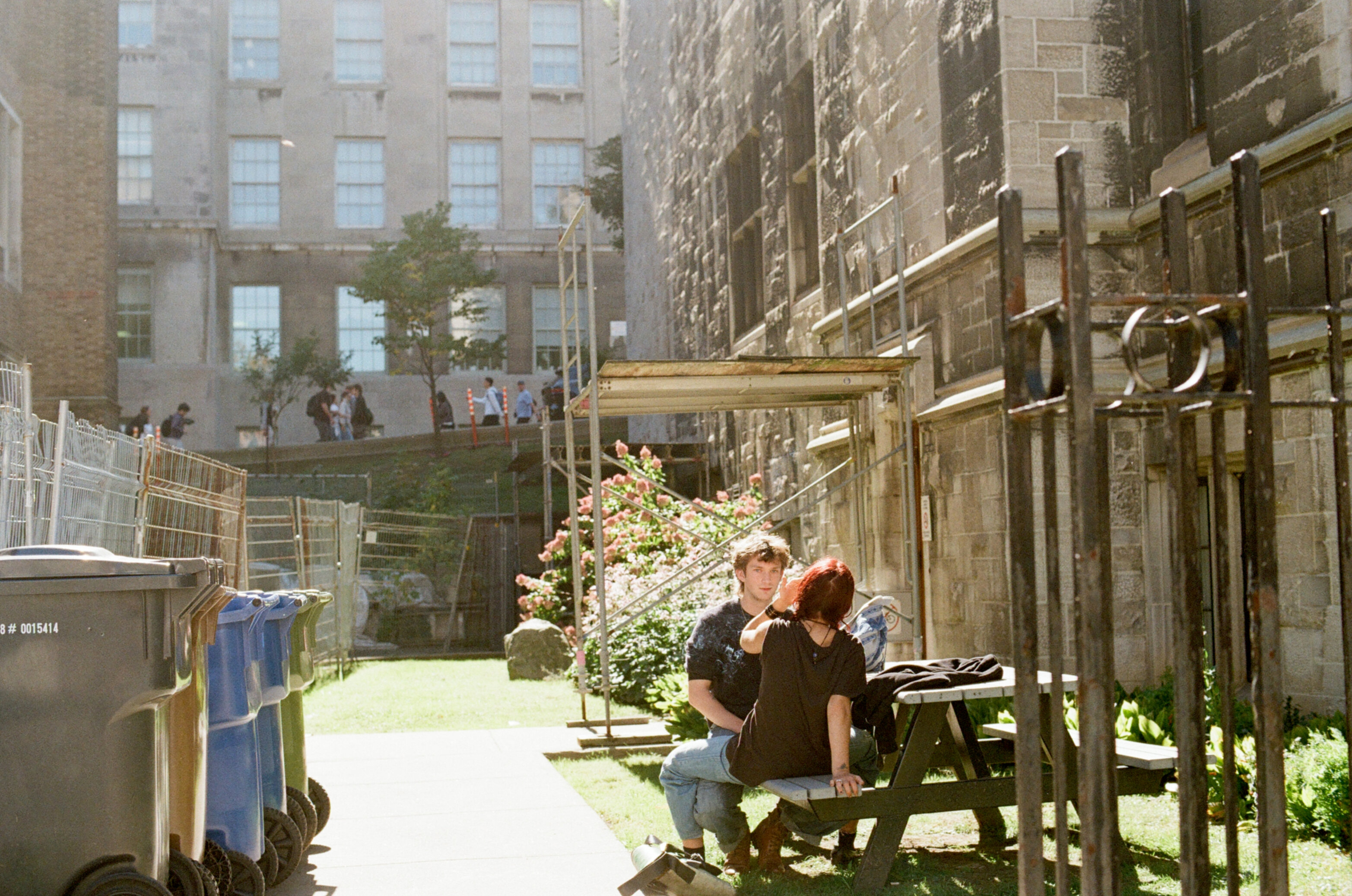 A male and female student sit next to each other, facing each other, at a picnic table in the sun next to a stone building
