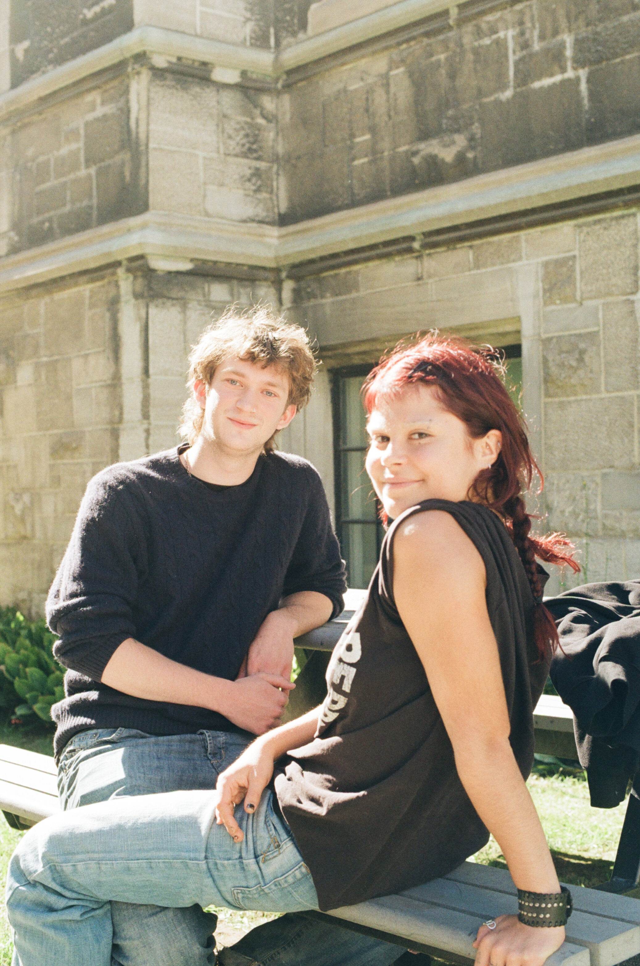 A male and female student sit next to each other, facing the camera, at a picnic table in the sun next to a stone building
