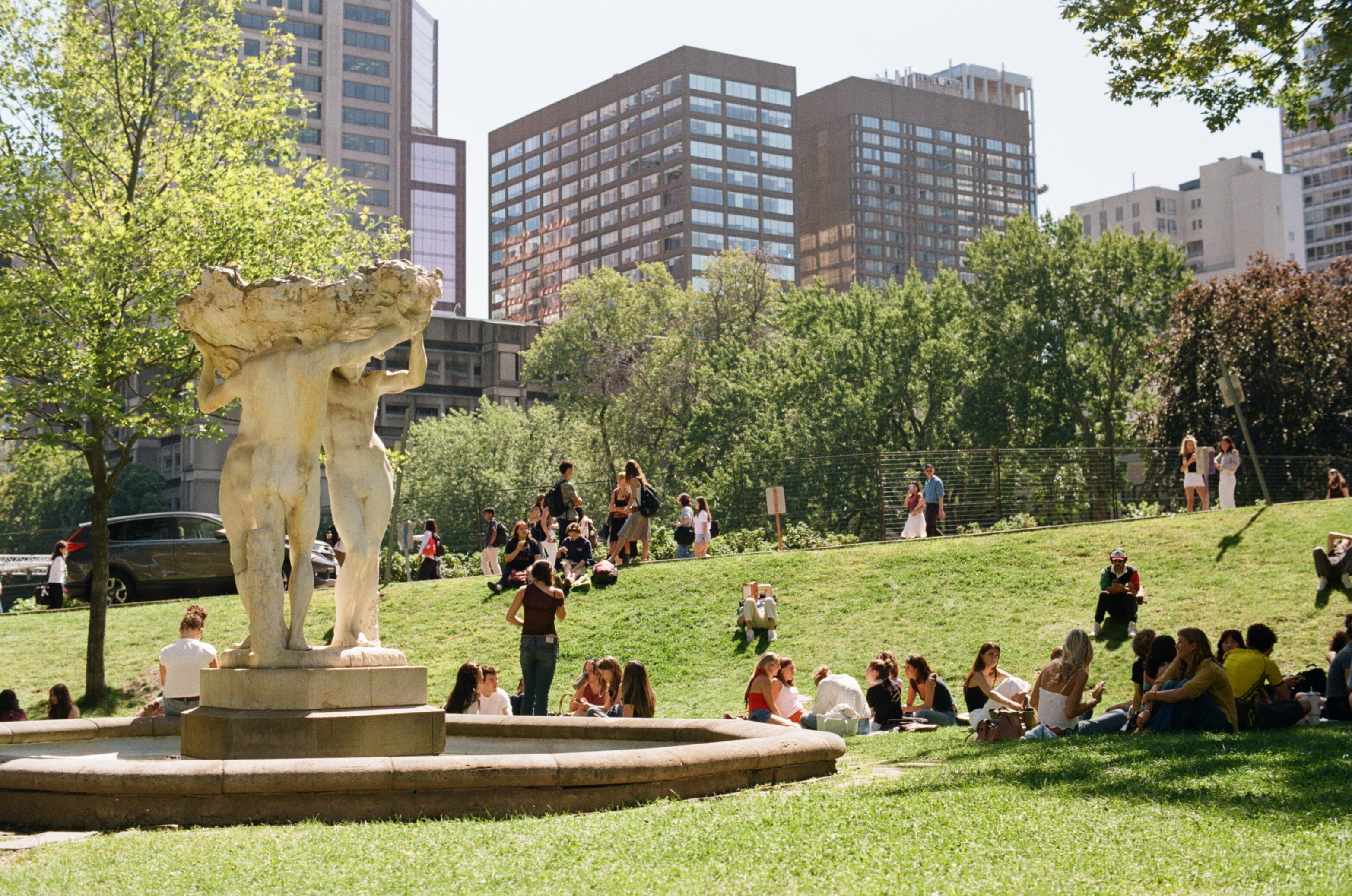 Students sitting on the grass next to the fountain with tall buildings in the background