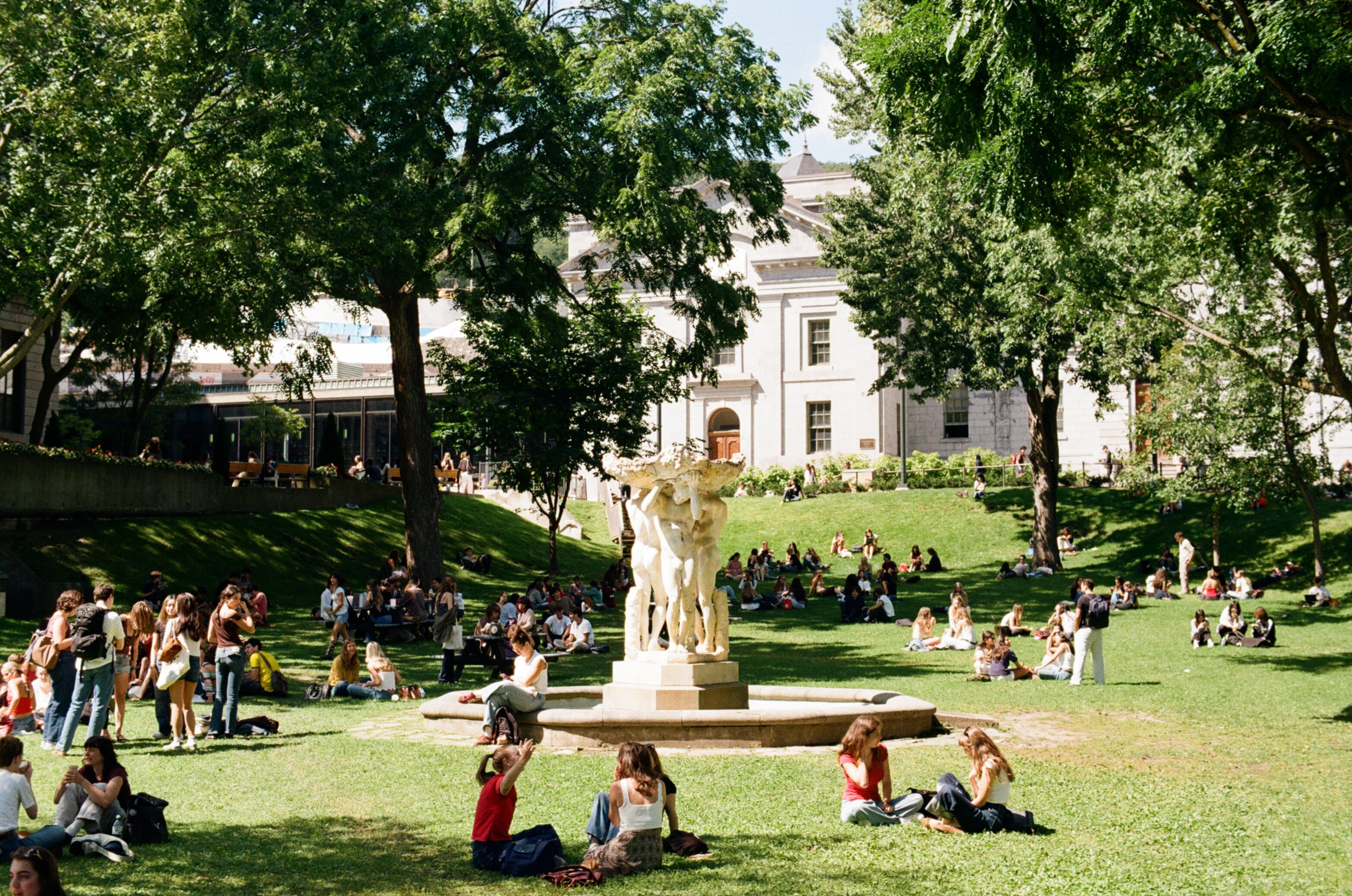 Students sitting on the grass around a fountain in the shape of three naked men