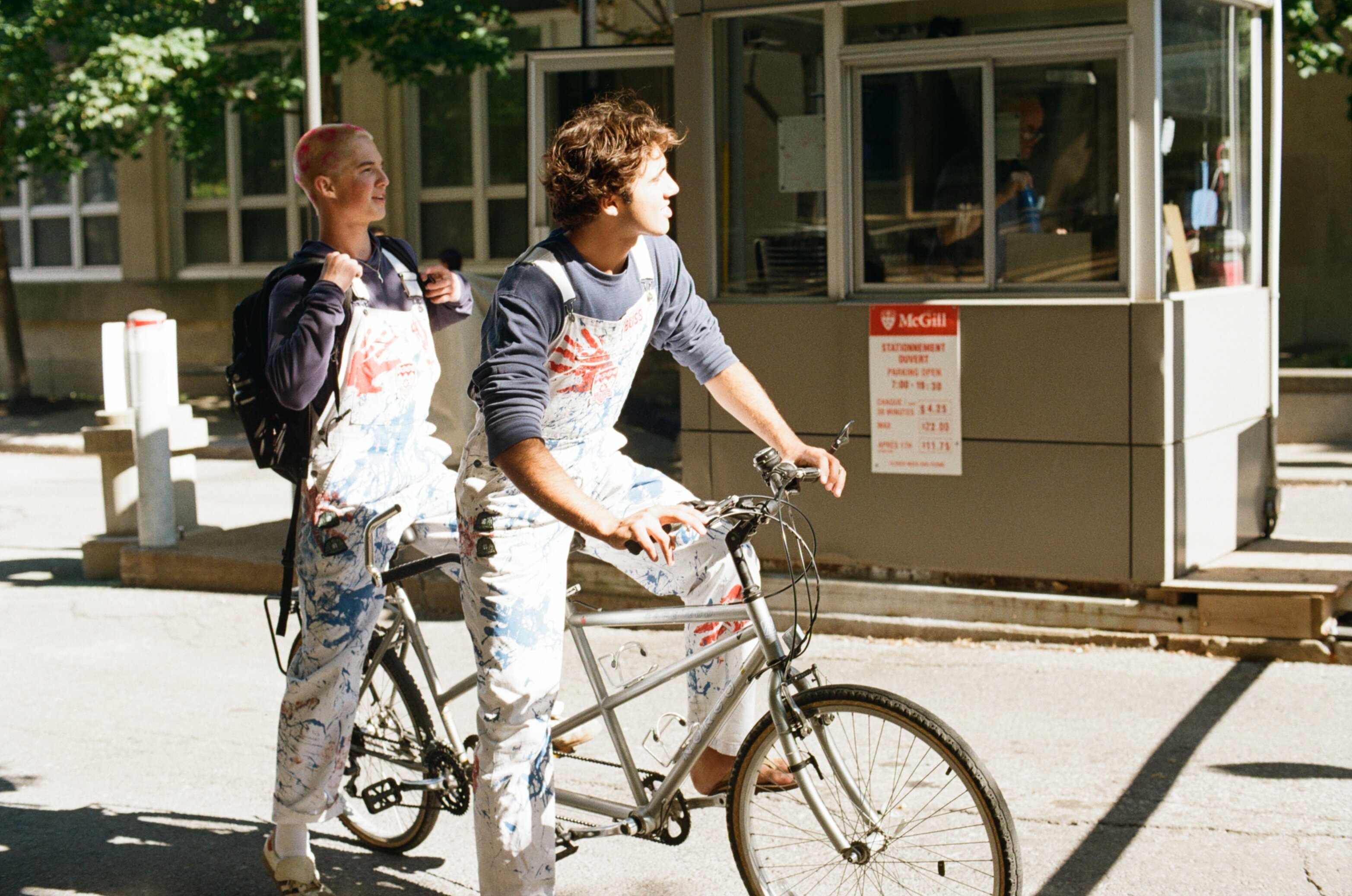 Two male students on a tandem bicycle wearing gray shirts and white overalls covered in splashes of colourful paint